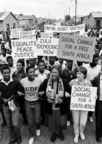 A historic black and white photo of a large crowd of people marching down a dirt street. They are holding signs with slogans like 'EQUALITY, PEACE, JUSTICE', 'ZULU DEMOCRACY NOW', and 'SOCIAL CHANGE FOR A FREE SOUTH AFRICA'.