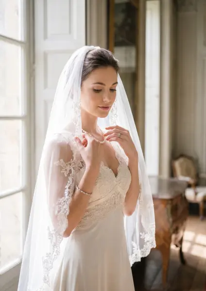 A serene young bride wearing a lace-trimmed wedding dress, gently holding the edges of her long, sheer veil near her face in a softly lit, elegant room.