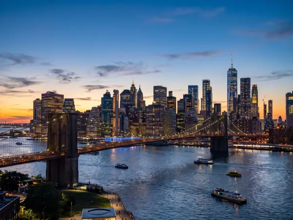 An aerial view showing the Brooklyn Bridge crossing the East River, with its suspension cables lit up. In the background, the dense skyline of Lower Manhattan is illuminated against a sky fading from sunset orange to twilight blue.