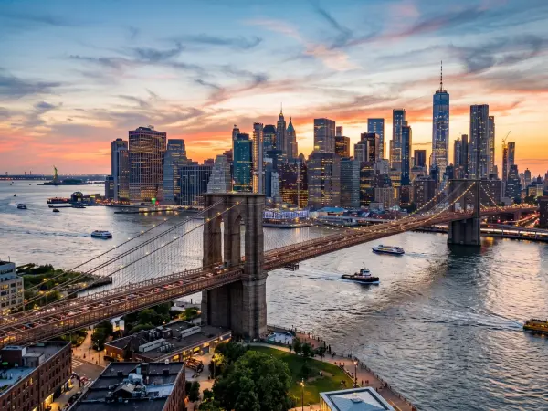 A breathtaking aerial shot of the Brooklyn Bridge spanning the East River at sunset. The sky is painted with vibrant orange and pink clouds, and the towering skyscrapers of Lower Manhattan, including One World Trade Center, glow in the background.