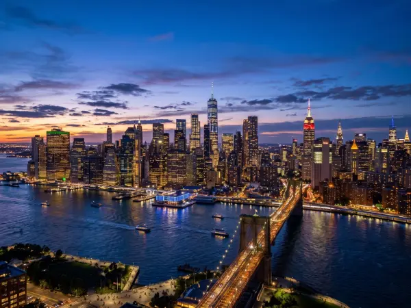 A stunning aerial view of the Brooklyn Bridge glowing with golden lights at dusk. Behind it, the dense skyline of Lower Manhattan is brilliantly illuminated against a deep blue evening sky, with ferries crossing the dark waters of the East River.