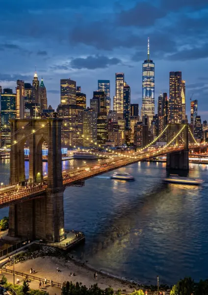 A high-angle view of the Brooklyn Bridge glowing with lights at dusk, spanning the East River. Behind it, the towering skyscrapers of the Financial District in Manhattan are brightly lit against a deep blue evening sky, with boats leaving light trails on 