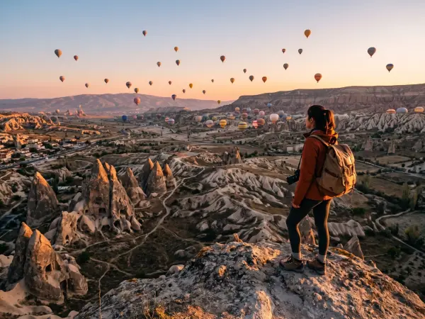 A woman wearing a red jacket and a backpack stands on a rocky overlook, holding a camera and gazing out at a vast valley filled with unique cone-shaped rock formations. The morning sky is filled with dozens of colorful hot air balloons illuminated by the 