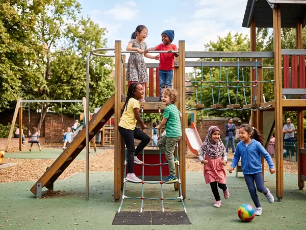 A lively scene of diverse children laughing and playing on a wooden climbing structure at an outdoor playground. Kids are climbing nets, running, and interacting joyfully on a sunny day.