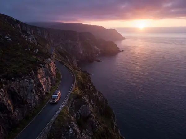 An aerial shot of a dark-colored SUV driving along a winding paved road that hugs the edge of a steep, rugged mountain cliff dropping off into the ocean. The scene is illuminated by a vibrant pink and orange sunset over the water.