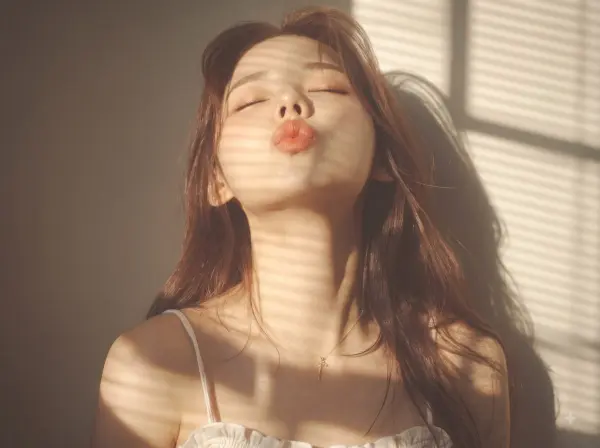 Woman with closed eyes blowing a kiss, face illuminated by sunlight with window blind shadows.