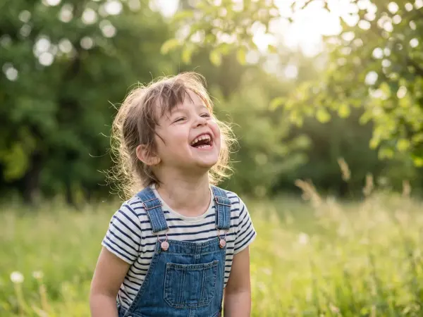 A close-up, heartwarming portrait of a cute little girl wearing a striped shirt and denim overalls. She is standing outdoors in a lush, green park, looking upwards and laughing with her mouth wide open, her hair softly backlit by the sun.