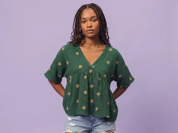 A portrait of a confident young woman with braided hair, wearing a loose green v-neck top with embroidered daisy flowers and distressed denim shorts, standing against a solid light purple background.