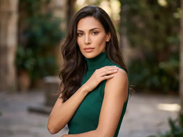 A close-up portrait of a strikingly beautiful woman with long, dark, wavy hair, looking directly at the camera with a gentle expression. She is wearing a fitted, dark green sleeveless turtleneck top, standing outdoors with a blurred stone courtyard in the