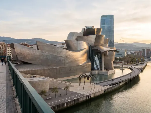 A breathtaking view of the Guggenheim Museum in Bilbao, Spain, bathed in the warm, golden light of sunset. The building's complex, curving titanium walls reflect the light. The famous giant spider sculpture, 'Maman', stands on the plaza bes