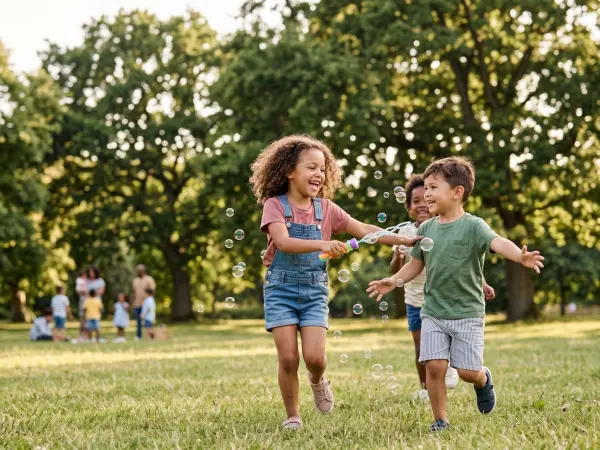 Three joyful, diverse young children are running towards the camera on a grassy lawn in a sunlit park. A girl in the front is holding a bubble wand, and they are all laughing and reaching out to catch the floating soap bubbles.