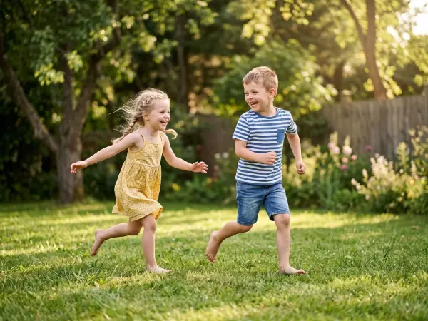 A happy little boy in a striped shirt and a little girl in a yellow dress are running barefoot across a lush green grassy lawn in a suburban backyard. They are both looking off to the side and laughing brightly on a sunny summer day.