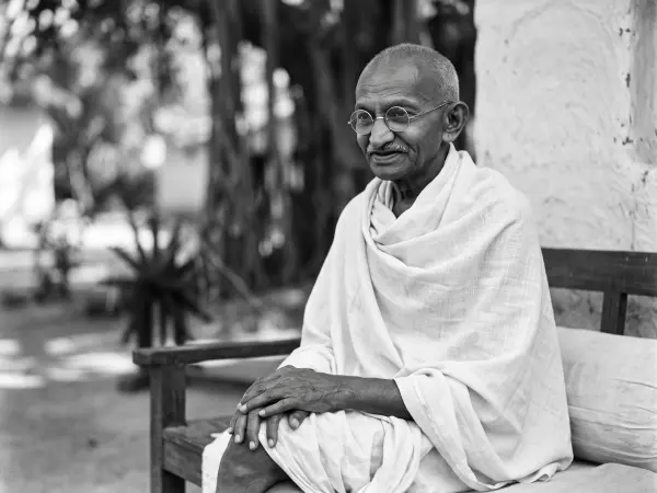 A black and white photograph of Mahatma Gandhi sitting on a wooden bench outdoors, wearing traditional white khadi cloth and round wire-rimmed glasses.