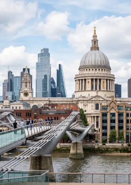 A scenic view of the Millennium Bridge in London spanning the River Thames, with several pedestrians walking across it. In the background stands the historic, domed St Paul's Cathedral, contrasting with the modern glass skyscrapers of the City of Lon