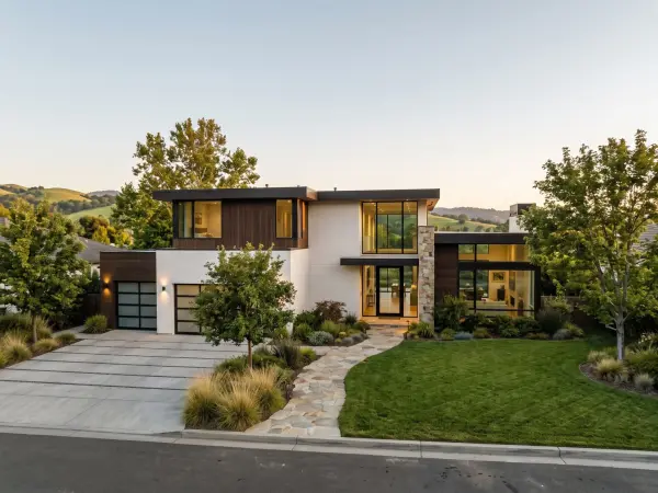 A large, two-story modern luxury home with white walls, dark wood paneling, and massive floor-to-ceiling windows glowing with warm interior light at dusk.