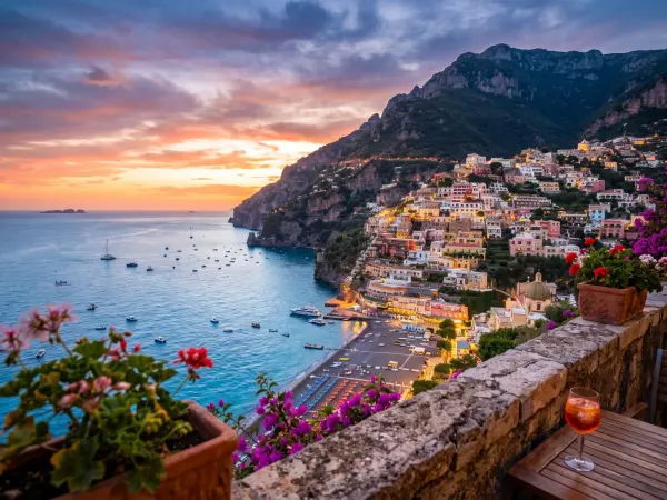 A stunning view of Positano, a colorful village built into a steep cliffside dropping down to a beach and the blue Mediterranean Sea, bathed in the light of a golden sunset. In the foreground is a stone terrace wall with bright pink flowers and a glass of
