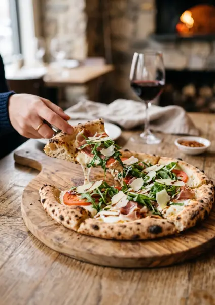 A hand lifting a slice of wood-fired pizza topped with prosciutto, arugula, tomatoes, and balsamic glaze, creating a gooey cheese pull, served on a rustic wooden board with a blurred fireplace in the background.