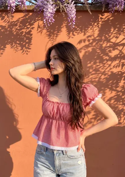 A beautiful young woman looking away with her hand in her hair, wearing a red and white gingham crop top and light blue jeans, standing against a sunlit orange wall beneath blooming purple wisteria flowers.