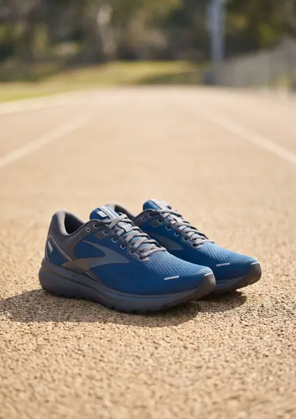 A pair of blue Brooks athletic running shoes with grey accents and white laces are sitting on the textured, reddish-brown surface of an outdoor athletic running track. The background is softly blurred showing the track lines fading into the distance.