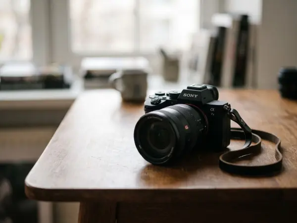 A close-up shot of a black Sony Alpha series mirrorless digital camera with a lens attached, sitting on a dark, rustic wooden desk. A dark leather camera strap is attached. The background is a bright, soft, out-of-focus room interior with a window and a c