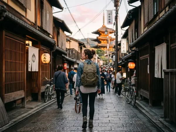A woman viewed from behind, wearing a denim jacket and a canvas backpack, walks down a narrow, cobblestone street lined with traditional Japanese wooden buildings and glowing paper lanterns. She holds a camera, and a tall pagoda is visible in the distance