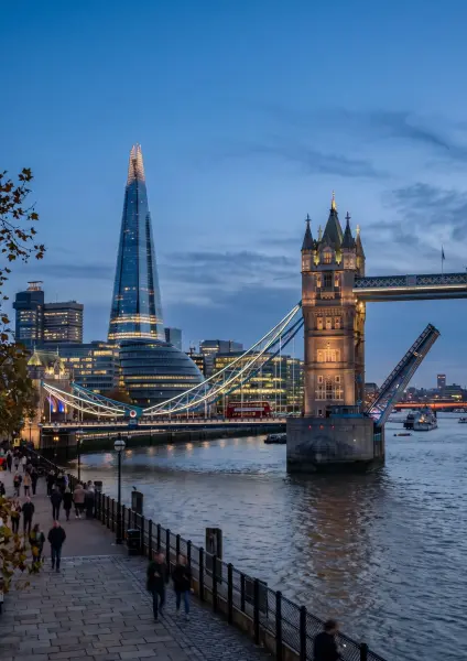 A beautiful twilight view of London showing the iconic Tower Bridge with its bascules raised to let a boat pass on the River Thames. To the left, the modern glass skyscraper known as The Shard is brightly illuminated against the darkening blue sky. Pedest