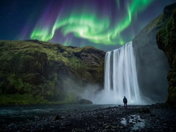 A silhouette of a person standing on a rocky shore looking up at a massive, powerful waterfall plunging from a mossy cliff. The night sky above is filled with swirling, vibrant green and purple aurora borealis (Northern Lights).