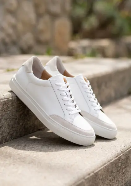 A close-up product shot of a pair of clean white leather sneakers with grey suede accents on the toe and heel. The shoes are resting casually on weathered outdoor stone steps, illuminated by bright sunlight with a blurred natural background.