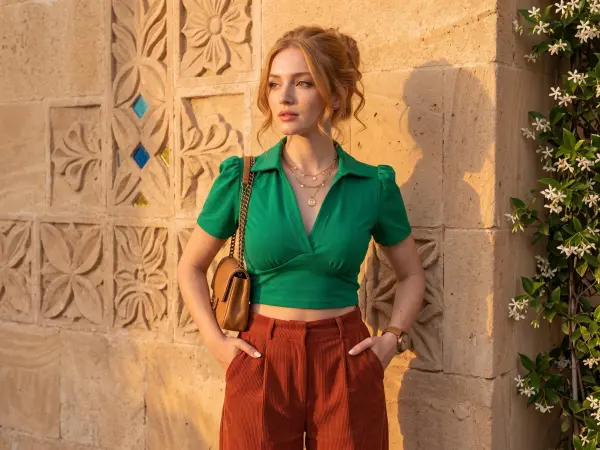 A beautiful young woman looking to the side, wearing a vibrant green v-neck short-sleeve top and rust-colored high-waisted corduroy pants. She has a brown leather bag on her shoulder and is posing against an intricately carved stone wall bathed in warm su
