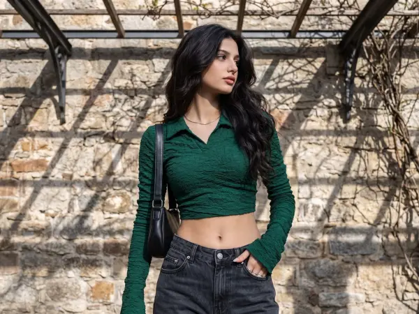 A beautiful young woman with long dark wavy hair is walking outdoors. She is wearing a dark green textured, long-sleeved crop top with a collar, dark grey jeans, and carrying a black shoulder bag, posing against a rustic stone wall.