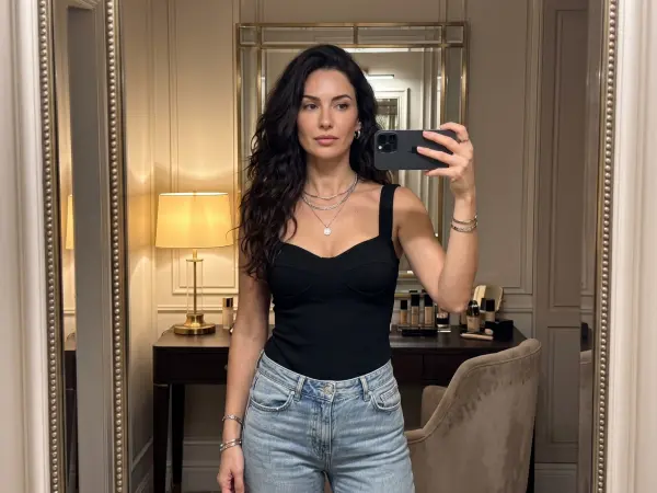 A beautiful woman with long dark wavy hair taking a selfie in a tall mirror using a black smartphone. She is wearing a fitted black bustier top and light wash blue jeans, standing in a bright, elegant room with a vanity desk in the background.