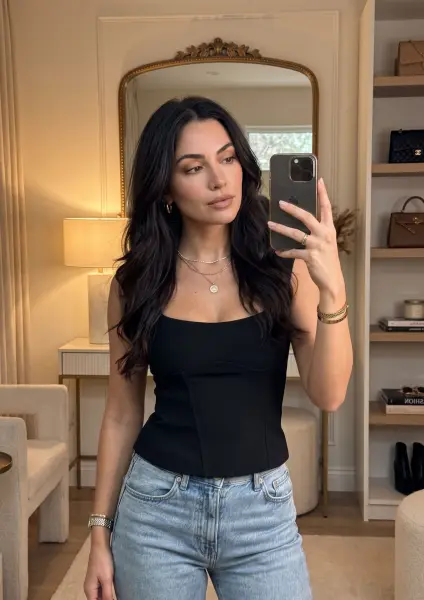 A beautiful woman with long dark hair holding an iPhone and taking a mirror selfie. She is wearing a black corset-style top and light blue jeans, standing in a chic, warm-toned room.