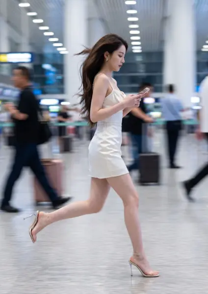 A young woman with long dark hair, wearing a short white dress and clear high heels, running quickly across the polished floor of a busy airport terminal while looking at her phone.