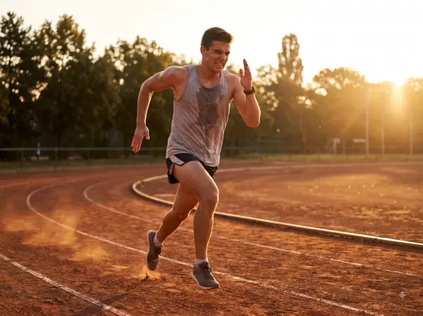 Sweaty athletic man with an intense expression sprinting on a red running track at sunset.